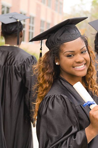 Candid moment of African descent female excitedly holding her diploma after the college graduation ceremony.  Her friends around her.  School building background on campus.